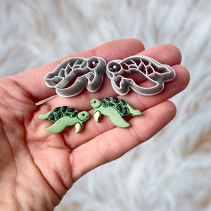 Könnte beinhalten: Zwei grüne Schildkröten-Anhänger aus Polymer Clay und zwei graue Kunststoff-Schildkröten-Ausstecher.