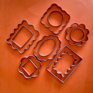 May include: A collection of five metal cookie cutters in various shapes, including a scalloped square, a scalloped rectangle, a scalloped circle, an oval with a scalloped edge, and a rectangular frame with a wavy inner border.