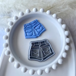 May include: A blue and grey pair of denim shorts cookie cutters. The cutters are on a white plate with a beaded edge.