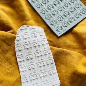 May include: Two white rectangular stamps with a repeating pattern of a vintage camper van design. One stamp is a solid block, the other is a sheet of stamps. The stamps are on a yellow-gold fabric. The text "Phoenix & Spruce" is visible on the stamp sheet.