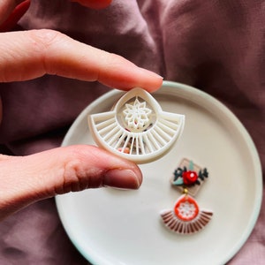 May include: White ceramic earring with a geometric design featuring a sunburst pattern and a flower in the centre. The earring is shown on a white plate with a second earring with a floral design.