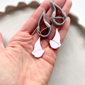 May include: A pair of gray and white gnome-shaped earrings. The earrings are made of polymer clay and have a matte finish.