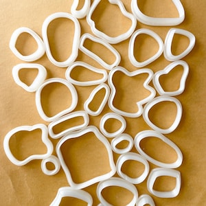 Könnte beinhalten: Ein Set mit 25 weißen Plastik-Ausstechern in verschiedenen organischen Formen, darunter Kreise, Ovale und freigeformte Formen. Die Ausstecher sind auf einer braunen Oberfläche angeordnet.