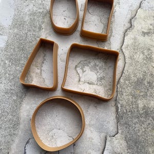 Puede incluir: Un conjunto de cinco cortadores de galletas de plástico marrón en varias formas geométricas: un óvalo, un rectángulo, un trapecio, un cuadrado y un círculo. Los cortadores están dispuestos sobre una superficie gris texturizada.