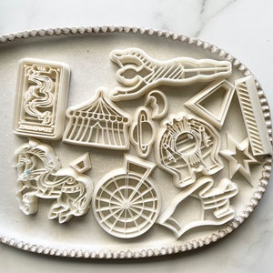 May include: A set of 12 white plastic cookie cutters in the shape of a circus theme. The cutters include a carousel horse, a tent, a star, a Ferris wheel, a clown, a crescent moon, a triangle, a striped bar, a tarot card, and a hand.