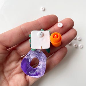 May include: A pair of colorful statement earrings with a purple, white, and green marbled design, a white square with a green leaf design, and an orange and white flower design. The earrings are attached to a silver post.