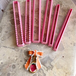Pu&ograve; includere: Tagliapasta rettangolari rosa con bordi smerlati e dritti sono disposti sopra un ciondolo a forma di fragola con un fiocco arancione. I tagliapasta sono di varie dimensioni, ideali per progetti di pasticceria e artigianato.