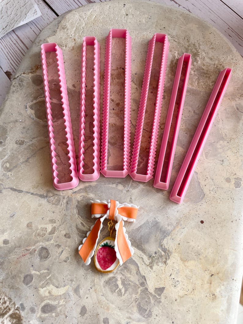 Pu&ograve; includere: Tagliapasta rettangolari rosa con bordi smerlati e dritti sono disposti sopra un ciondolo a forma di fragola con un fiocco arancione. I tagliapasta sono di varie dimensioni, ideali per progetti di pasticceria e artigianato.