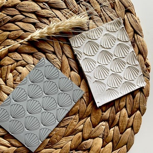 Könnte beinhalten: Zwei graue und weiße Gummistempel mit einem Muschelmuster. Die Stempel liegen auf einer geflochtenen braunen Matte.