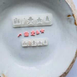 Puede incluir: Un juego de moldes de silicona transparente para hacer decoraciones con temática navideña. Los moldes están dispuestos en dos filas, con la fila superior que muestra una caja de regalo, un bastón de caramelo, un hombre de jengibre, una estrella y un árbol de Navidad. La fila inferior muestra un bastón de caramelo, una caja de regalo, un hombre de jengibre, un árbol de Navidad y una estrella. Los moldes están llenos de arcilla polimérica rosa.