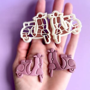 K&ouml;nnte beinhalten: Zwei wei&szlig;e Plastik-Ausstecher in Form von Vintage-Rollern und zwei lila Fondant-Kekse in Form von Vintage-Rollern.