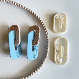 May include: Two pairs of blue and brown clay earrings with floral designs. The earrings are on a white plate with two white plastic molds.