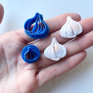 Puede incluir: Una mano sostiene un cortador de galletas azul con forma de ajo y dos adornos blancos con forma de ajo. El cortador de galletas es de plástico azul. Los adornos son blancos con detalles marrones y negros, que se asemejan a bulbos de ajo reales. Los artículos se muestran sobre una superficie clara.