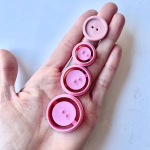 Peut inclure: Quatre boutons roses de différentes tailles sont présentés. Les boutons sont ronds avec deux ou quatre trous. Les boutons sont d'une couleur rose clair unie. Les boutons sont disposés en ligne verticale.