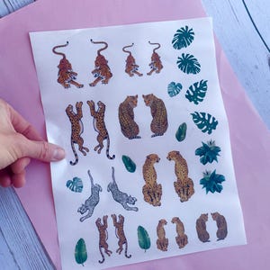 Puede incluir: Una hoja de pegatinas con temática de animales con tigres, leopardos y hojas tropicales en tonos naranja, marrón y verde. Las pegatinas están dispuestas sobre un fondo blanco, sujetas sobre una superficie rosa.