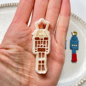 Pode incluir: Cortador de biscoitos de pl&aacute;stico branco em forma de quebra-nozes. O cortador tem um design detalhado com um chap&eacute;u, um rosto e um corpo.