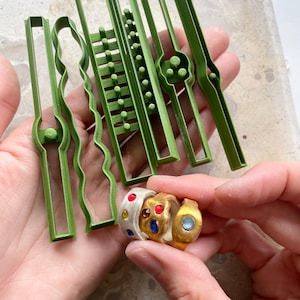 Può includere: Una collezione di strumenti verdi a forma di anello e tre anelli colorati. Gli anelli sono in oro e argento, adornati con pietre preziose rosse, blu e gialle. Gli strumenti hanno varie forme, tra cui linee rette, ondulate e design a scala.