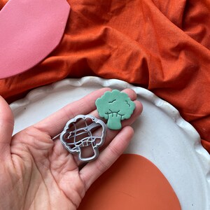 May include: A hand holds a green broccoli-shaped clay piece and a silver metal cookie cutter. A pink and orange clay piece and a white plate are also visible. The background is an orange fabric.