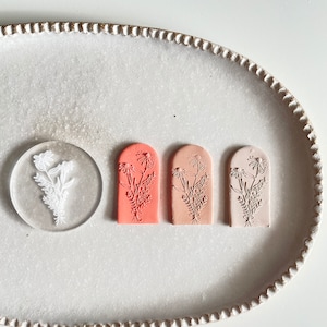 May include: Three arch-shaped clay stamps with a floral design. The stamps are in shades of pink, peach, and white. The stamps are arranged in a row on a white plate.