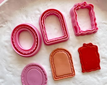 Può includere: Sei tagliabiscotti in plastica rosa e rossa a forma di cornici. I tagliabiscotti sono su una superficie bianca.