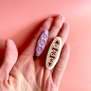 May include: Two surfboard-shaped cookie cutters. One is lilac, the other is off-white. Each cutter has three flower-shaped cutouts. The cutters are held in a hand against a pink background. The cutters are likely for making biscuits or fondant decorations.