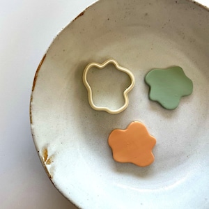 May include: Three clay shapes on a white plate. The shapes are a green cloud, an orange cloud, and a white cookie cutter in the shape of a flower.