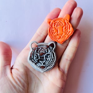 May include: Two tiger head-shaped cookie cutters. One is orange, the other is silver. The orange cutter has a detailed tiger face design. The silver cutter has a detailed tiger face design with black stripes. Both are held in a hand.