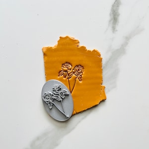 May include: A close-up of a grey polymer clay stamp with a floral design. The stamp is on a yellow surface.