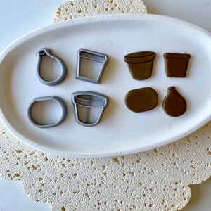 May include: A set of eight cookie cutters in various shapes, including a vase, a pot, a flower, and a leaf. The cutters are made of silver-coloured metal and are arranged on a white plate.