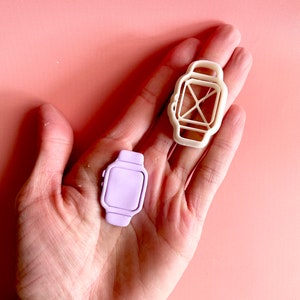 May include: Two plastic cookie cutters in the shape of a smartwatch. One cutter is white and the other is light purple.
