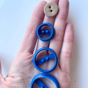 May include: A selection of sewing supplies, featuring a wooden button with two holes and three blue plastic button-shaped moulds in different sizes. The moulds are circular with a bar and two raised dots.