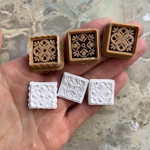 May include: A hand holds six small, square decorative tiles. Three are tan with a dark brown floral design, and three are white with a raised floral pattern. The tiles are likely for crafts or home decor.