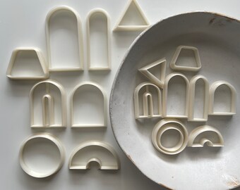 May include: A set of white plastic cookie cutters in various shapes, including arches, circles, semicircles, and triangles. The cutters are arranged on a white surface and in a white ceramic bowl.