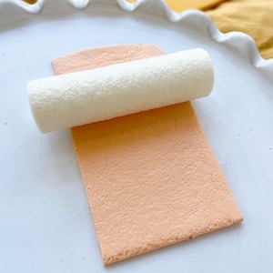 May include: A white rolling pin is rolling over a light peach-coloured fondant sheet on a white plate.