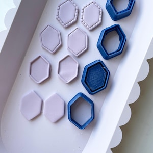 Puede incluir: Una bandeja blanca con un borde festoneado contiene varios cortadores de galletas hexagonales y trozos de arcilla morada. Los cortadores de galletas son de plástico azul.