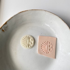 May include: Two white rubber stamps with a sun and star design on a white ceramic plate. The stamps are circular and have a raised design.