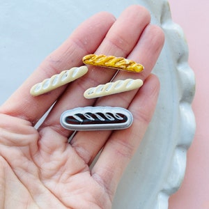 Puede incluir: Cuatro amuletos de baguette de pan en miniatura en varios tonos de blanco, amarillo y marrón. Los amuletos están hechos de arcilla polimérica y tienen un aspecto realista.