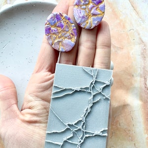 Puede incluir: Dos pendientes ovalados de color morado y dorado con textura craquelada. Un molde de silicona rectangular gris con textura craquelada.