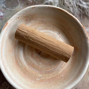 Può includere: Un pezzo cilindrico di legno marrone chiaro con una visibile trama a venature verticali, appoggiato all'interno di una ciotola in ceramica rotonda e poco profonda. La ciotola presenta una base color crema con sottili macchie vorticose color terracotta.