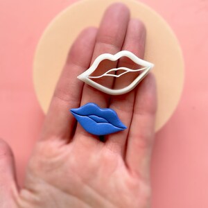 May include: Two cookie cutters in the shape of lips. The larger cutter is white and the smaller cutter is blue.