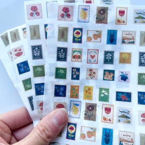 Puede incluir: Hojas de pegatinas decorativas con varios diseños que se asemejan a sellos postales. Los sellos muestran ilustraciones florales, frutales e insectos en una variedad de colores, incluyendo rojo, azul y amarillo. Las pegatinas se sostienen en una mano.