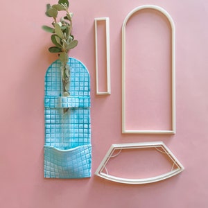 May include: A blue ceramic wall vase with a small glass tube for flowers. The vase has a square tile pattern and a pocket on the bottom. The vase is surrounded by white plastic stencils in the shape of an arch, a rectangle, and a curved shape.