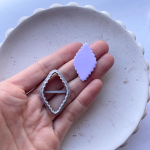 Puede incluir: Un cortador de galletas de metal plateado en forma de diamante con bordes festoneados. También se muestra una forma de arcilla polimérica de color púrpura claro.