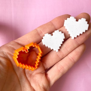 May include: Three pixelated heart shapes, one orange and two white, are shown in a hand. The orange heart is a cookie cutter, and the white hearts are made of a textured material.