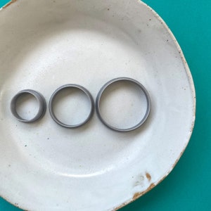 Puede incluir: Tres cortadores de galletas de metal gris de diferentes tamaños en un plato de cerámica blanco. El plato tiene un esmalte moteado y un borde marrón.