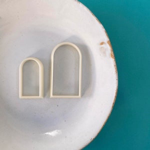 Op de afbeelding: Twee witte plastic koekjesvormen in de vorm van boogvensters, een groter dan de andere, op een wit bord met een gespikkeld oppervlak.