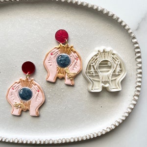 May include: Two pink clay earrings with gold accents and a blue and white circle in the center. The earrings are on a white plate with a scalloped edge. A white plastic mold of the earrings is also on the plate.