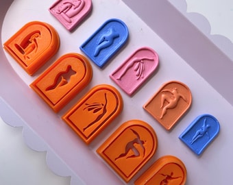 May include: A set of nine arch-shaped silicone molds featuring various female body shapes in orange, pink, and blue. The molds are arranged on a light purple tray with a scalloped edge.