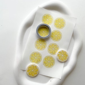 Może przedstawiać: Arkusz papieru z dziewięcioma żółtymi naklejkami w kształcie plastrów cytryny. Jedna naklejka znajduje się na rolce taśmy.