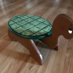 May include: A wooden turtle stool with a green painted shell and brown painted body. The stool has a rounded top and four legs.